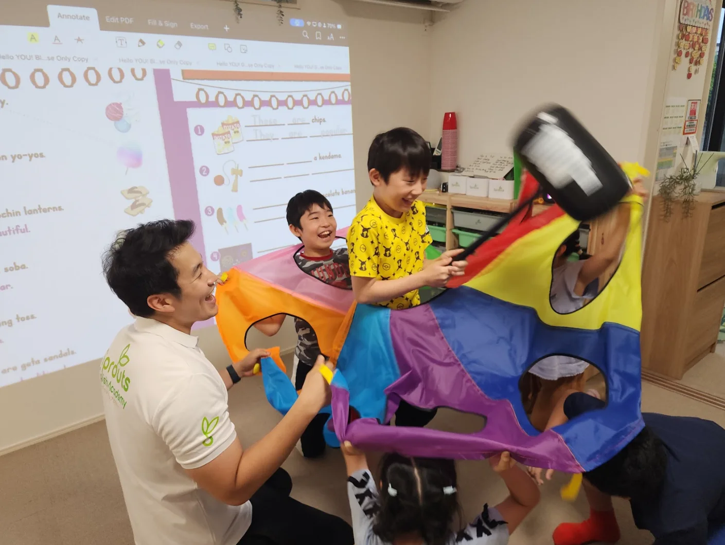 先生と子どもたちがパラシュート遊びのような活動をしながら、楽しく英語を使っている様子